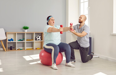 Plus size, fat woman training with dumbbells under guidance of personal trainer in gym. Obese, overweight woman sitting on fitball doing exercise with dumbbells. Weight loss, healthy lifestyle
