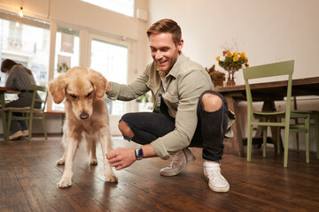 Portrait of happy man spending time with his dog in pet-friendly cafe, playing and petting golden...