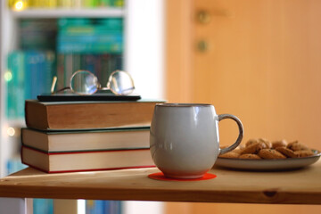 Cup of tea or coffee, pile of books, plate of cookies, reading glasses, e-reader and pen on the table. Colorful rainbow bookshelf in the background. Selective focus.