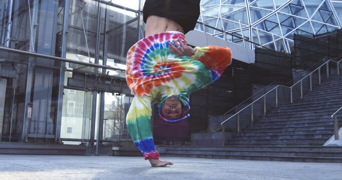 Super slow motion of young modern male break dancer in colorful urban street style wear is performing energetic choreography with backflip in the middle of urban city center streets.