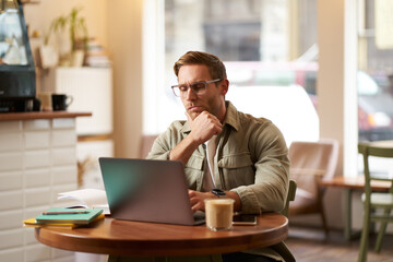Portrait of businessman in glasses, sits in cafe with laptop, looks concentrated at screen with his...
