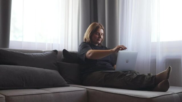 Happy young caucasian woman using texting mobile phone or chat and smiling with laptop computer sitting of the sofa in home. Communication female looking message at cell phone or smartphone. Slow