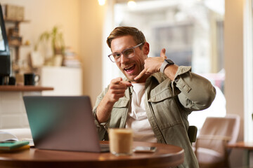 Portrait of cheerful, flirty guy in glasses, sitting with laptop computer in cafe, showing phone call hand gesture and winking at you, giving his number - obrazy, fototapety, plakaty