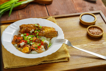 Thai chicken with rice and vegetables in a white plate on a wooden table next to soy sauce and vegetables in the background