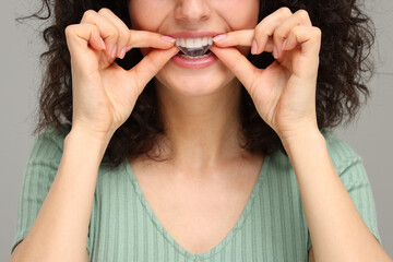 Young woman applying whitening strip on her teeth against grey background, closeup