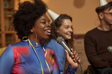 Side view portrait of Black young woman singing to microphone and enjoying karaoke during home...