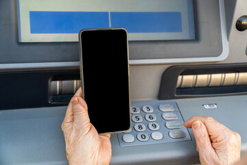 Older woman hands with an smartphone close to an ATM.