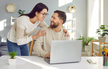 Happy young idyllic family couple using their laptop computer together. Loving woman smiling and holding her husband by the shoulders while he is sitting at his working desk with a notebook PC