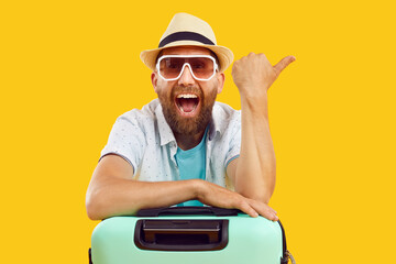 Portrait of young happy funny man tourist in sunglasses and beach hat with suitcase pointing index...