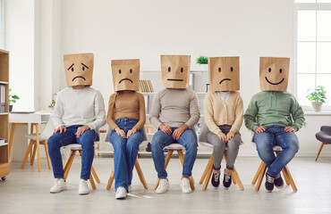 Human emotions. Portrait of group of unrecognizable people wearing paper bags on their heads with different emotions drawn on them. Men and women sit in row on chairs and express different emotions.  - 765126985