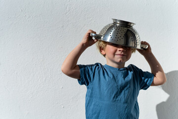 Little boy putting colander on his head . cheerful blond boy with a sieve on his head. positive kid - obrazy, fototapety, plakaty