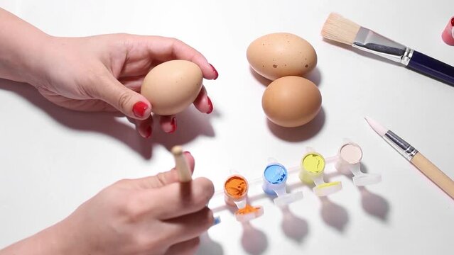 White woman paints letter A on beige chicken egg with blue paint. Female hand with red manicure. Happy Easter concept, preparation for religious holiday, religion, art. Brushes on white background