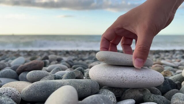 Female hand builds pyramid of light stones on pebble beach. Ocean waves. White woman experiences Zen on sea shore. Concept of spa, calmness, yoga, self-improvement, construction, games, travel