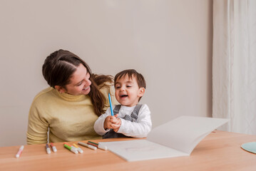 Fototapeta premium Happy Young toddler Boy Drawing with Markers sitting on the table with his smiling mother at Home