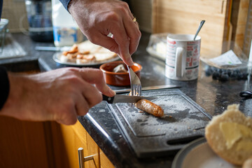 Male in the kitchen making tea and cutting sausage with knife and fork. 