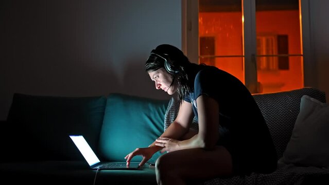 Woman Uses Laptop At Night In Living Room - Sitting On Sofa With Lights Off And Headphones On