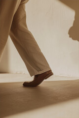 Close-up of a person's leg in white pants and brown shoes walking on a white surface, with soft light and shadows. 

