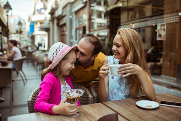 Family enjoying time at a cafe terrace with father kissing daughter and mother having coffee