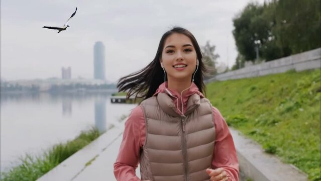 J&oacute;ven bonita trotando por la ma&ntilde;ana a orillas del r&iacute;o, mientras escucha m&uacute;sica con sus auriculares, viste una blusa rosa pastel y una campera marr&oacute;n. Un ave vuela a su lado acompa&ntilde;&aacute;ndola.