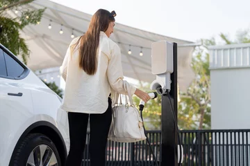 Foto op Canvas Young woman holding shopping bag recharge EV car battery from charging station at parking lot. Modern woman go shopping by environmental friendly electric vehicle in urban travel lifestyle. Expedient © Summit Art Creations