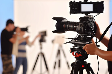 A camera on a tripod being used to film a scene. There are also two photographers in the background, preparing to take photos.