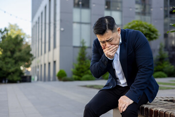 Obraz premium Asian young man in a suit sits on a bench in the street with his hand covering his mouth, feels severe nausea and urge to vomit