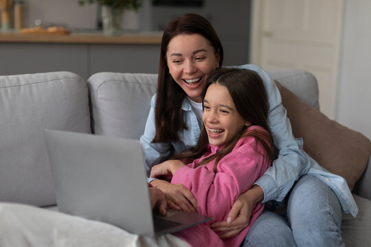 Happy woman hugging laughing daughter while using laptop, surfing internet or watching film together on sofa at home