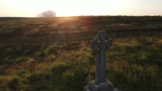 A drone shot of a celtic cross with a sunset and smoke from a gorse fire in the background. Bragan, Monaghan, Ireland