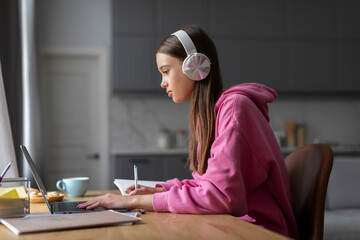 Focused teen girl wearing headphones studying online with laptop from home, making notes, doing...
