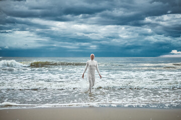 Hairless girl with alopecia in white futuristic suit comes out of cold restless sea on sandy beach,...