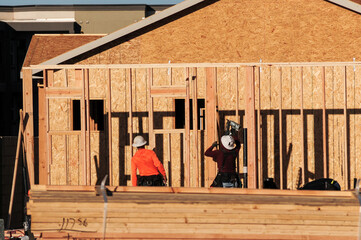 In a housing development in Queen Creek, Arizona, two wood framers wearing hard hats are using power equipment, such as a pneumatic nailer, to construct the wooden framework of a tract home - obrazy, fototapety, plakaty