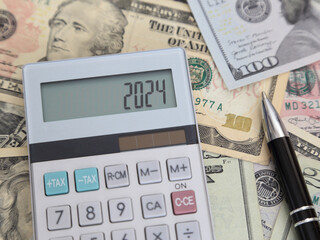 2024 is shown on a calculator LCD screen among multiple denominations of USA dollar bills and next to an ink pen, demonstrating finance, business, and budget related concepts.