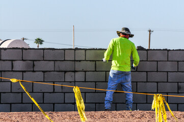 A construction worker applies mortar to a concrete block wall at a marked job site, depicting...