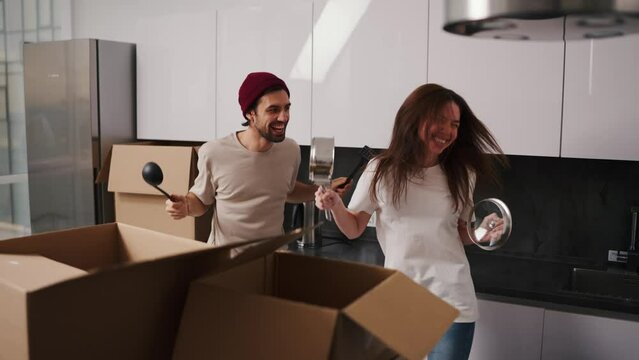 A happy brunette girl in a white T-shirt and a guy in a red hat and beige T-shirt are dancing holding kitchen utensils in their hands during their move to a new apartment they are happy and joyful