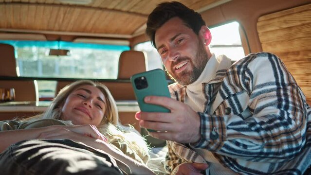A cozy scene inside a campervan where a man and woman lie on a bed, smiling and sharing a moment over a smartphone.