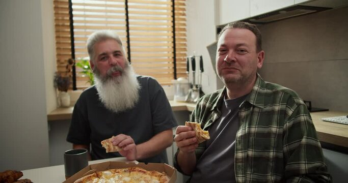 Portrait of a happy LGBT couple of two men brunette in a checkered shirt and an older man with gray hair and a lush gray beard who eat pizza in the kitchen during their lunch and are happy