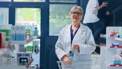 Aged knowledgeable druggist in pharmacy holding shopping basket, arranging merchandise on shelves....