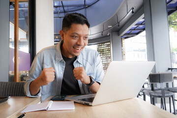 Asian man happy and enthusiastic with blue shirt using laptop and mobile phone in coffee shop,...