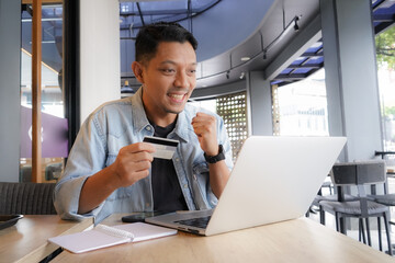 Asian man using laptop and mobile phone for shopping online with credit card in coffee shop online...