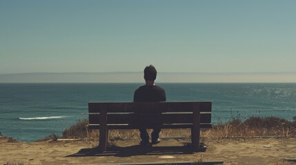 A man sitting on a bench staring out at the ocean with a sad expression as he contemplates his upcoming relocation. - obrazy, fototapety, plakaty