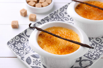 Delicious creme brulee in bowls, vanilla pods and sugar cubes on white wooden table, closeup