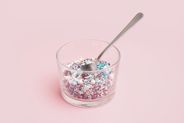 Glass and spoon with sweet colorful sprinkles on pink background