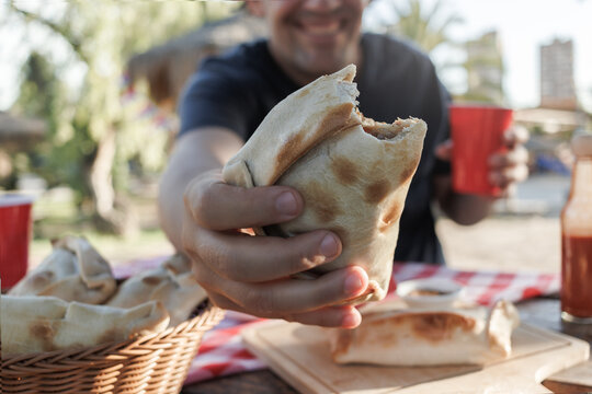 Unrecognizable Latino man laughing and showing a bitten Chilean pino empanada. Sitting at wooden table in park, with more empanadas in wicker basket. Park vegetation in the background