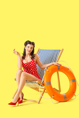 Beautiful pin-up lifeguard with ring buoy and whistle sitting on deck chair against yellow background
