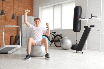Sporty young man with dumbbells training on fitball in gym