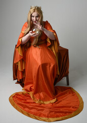 Full length portrait of plus size blonde woman, wearing historical medieval fantasy gown, golden crown  royal queen. sitting pose on throne chair, isolated studio background.