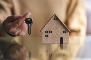 Closeup image of a woman holding the keys with a wooden house model on the table for real estate concept