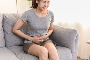 Healthcare medical or daily life concept : Close up hands of young Asian woman holding stomach have a stomachache or menstruation pain sitting on her sofa.