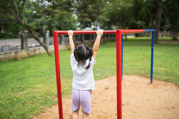 A little girl is hanging from a red and blue park bar. She is wearing a white shirt and purple shorts
