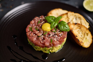 Tuna and avocado tartare with sesame seeds, capers and egg yolk on a dark ceramic plate with baguette bread crouton chips